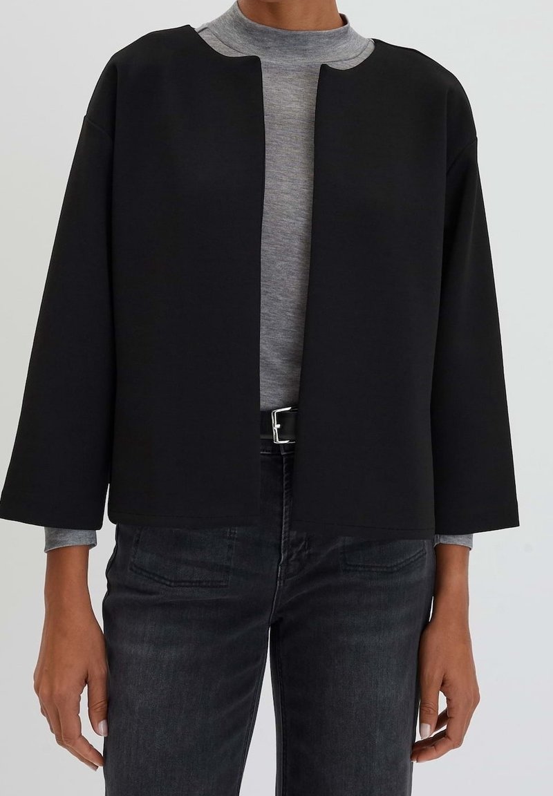 Svart kort jacka med en fyrkantig form, vida ärmar och öppen front. Bärs över en grå höghalshörning, i kombination med mörka jeans.