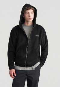 Sweat à capuche noir zippé en tissu texturé et doux. Comprend une capuche, des poches avant et une petite étiquette sur la poitrine. Porté par-dessus une chemise grise.