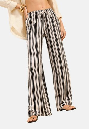 Femeie purtând pantaloni cu talie înaltă și largi, cu dungi verticale negre și bej, asortați cu o cămașă deschisă bej și sandale joase.