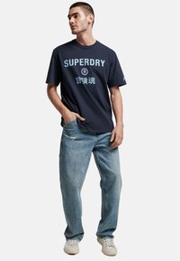 T-shirt in cotone blu navy con logo "SUPERDRY" in azzurro, abbinato a jeans chiari strappati e sneakers bianche.
