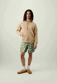 Sweatshirt bege com um bolso frontal, calções verdes com um padrão de folhas e sapatos slip-on bege com meias brancas. Design simples e cores neutras.