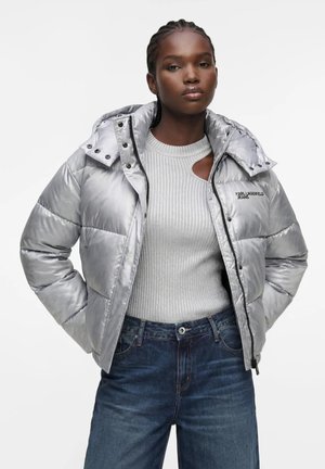 Ung kvinde står med hænderne i lommerne iført en skinnende sølv pufferjakke, hvid ribstrikket sweater og blå denimjeans.