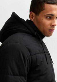 Chaqueta de plumas negra con capucha, que presenta una textura acolchada y cordones ajustables. Tejido suave y forma aerodinámica.