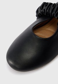 Zwarte leren ronde platte schoen met geconfigureerde elastische opening en tan-kleurige gevoerde binnenzool op een witte achtergrond.