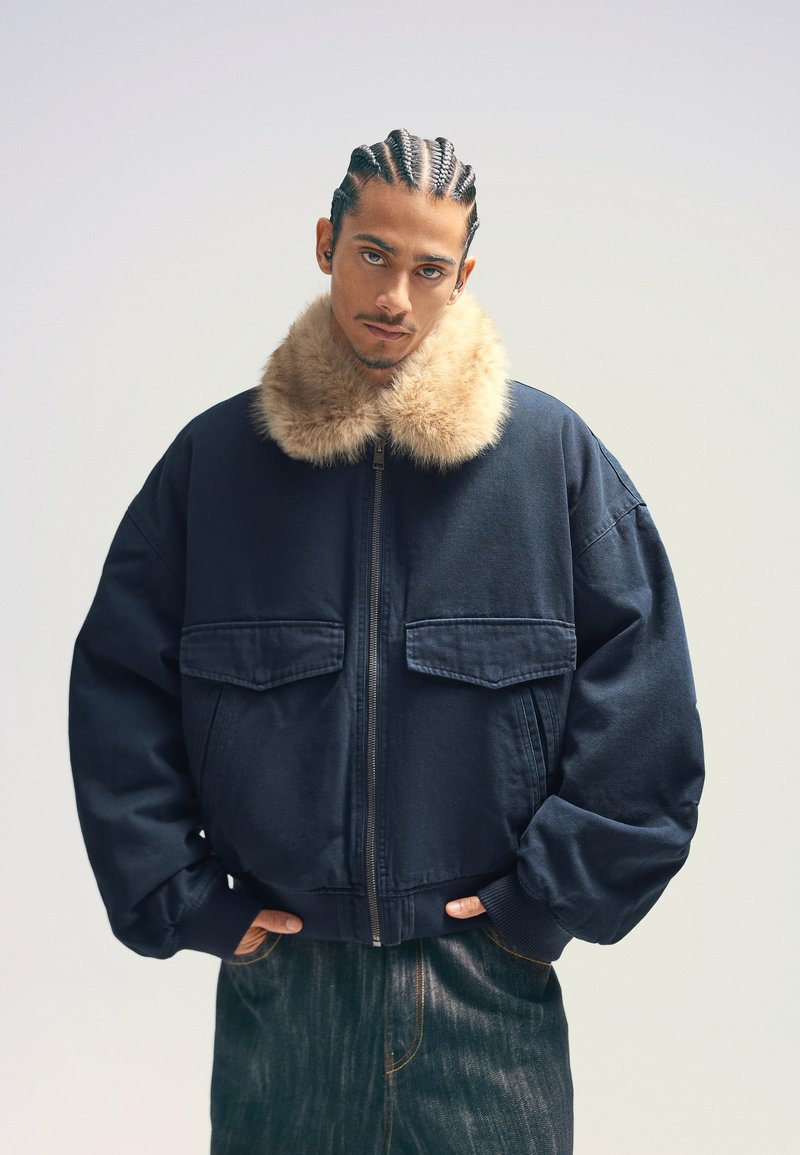 Marinblå oversizejacka med furfoder på kragen, framfäste med dragkedja och två bröstfickor, kombinerad med mörka denimbyxor.