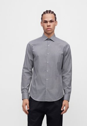 Jeune homme aux cheveux tressés portant une chemise gris à carreaux boutonnée et un pantalon noir, debout devant un fond clair uni.