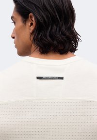 Άνδρας με σκουρόχρωμα μαλλιά έως τους ώμους φοράει λευκό υφασμάτινο πουκάμισο με μαύρη ετικέτα που γράφει «All Conditions Gear» στο πάνω μέρος της πλάτης.