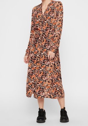 Žena v midi šatech s dlouhým rukávem s oranžovo-růžovým květinovým vzorem, doplněná černými teniskami, stojící před jednoduchým pozadím.