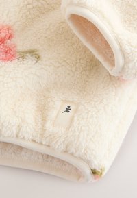 Tela de forro polar de color crema suave con superficie texturizada, un pequeño símbolo de hoja negra en una etiqueta cosida y sutiles detalles de color.