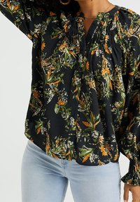 Květinová blůza s černým pozadím, zdobená oranžovými a zelenými vzory, s nařaseným výstřihem, detaily na knoflíky a dlouhými rukávy.