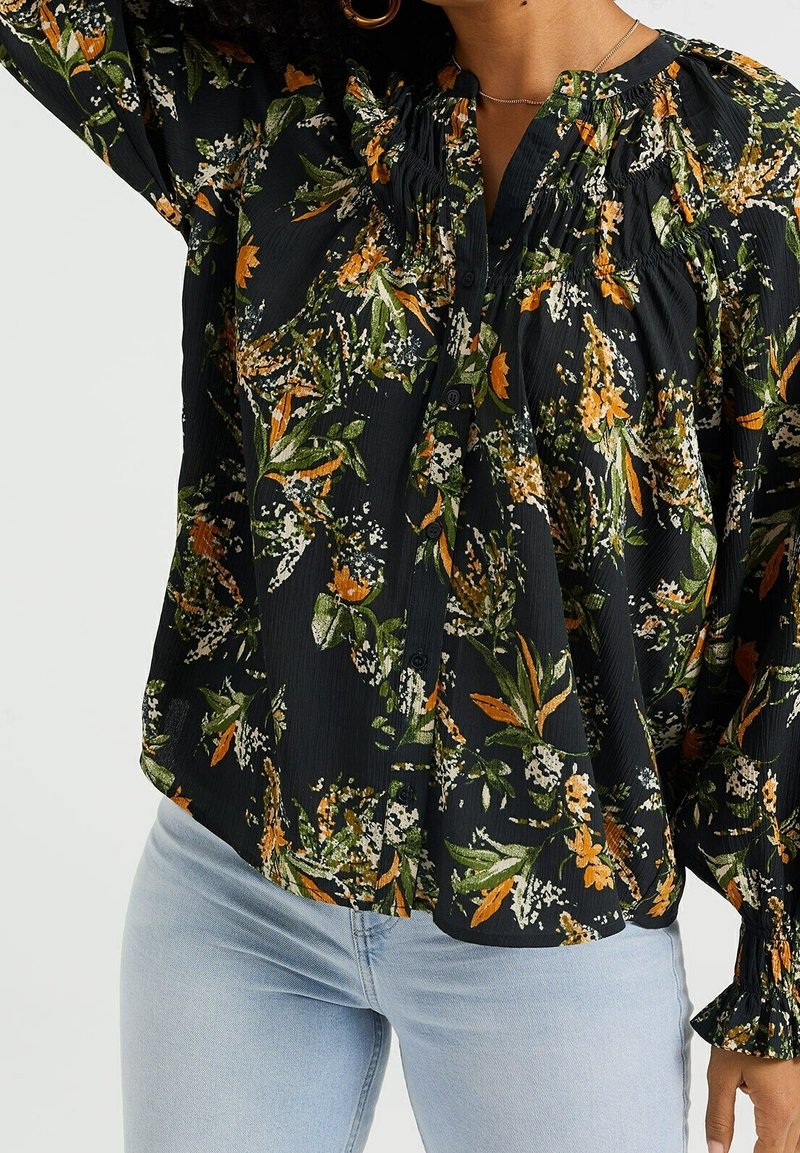 Květinová blůza s černým pozadím, zdobená oranžovými a zelenými vzory, s nařaseným výstřihem, detaily na knoflíky a dlouhými rukávy.