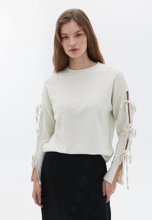 Maglione di colore crema con texture, dotato di fessure laterali e dettagli annodati sulle maniche. Indossato sopra una gonna nera, presentato su uno sfondo minimalista.