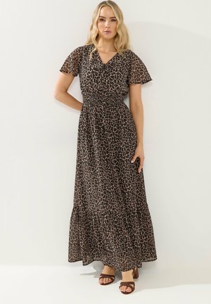 Kvinde iført en maxi-kjole med brunt og sort leopardmønster, korte flagrende ærmer og brune hælede sandaler, stående mod en hvid væg.