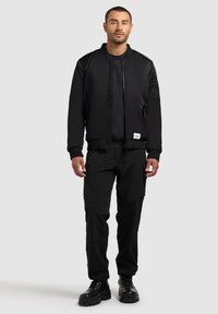 Giubbotto bomber nero con zip frontale, polsini a coste e tasche laterali, abbinato a pantaloni cargo neri e stivali neri, tutti in un design elegante.
