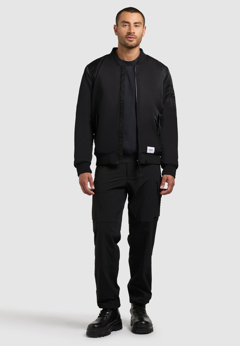 Giubbotto bomber nero con zip frontale, polsini a coste e tasche laterali, abbinato a pantaloni cargo neri e stivali neri, tutti in un design elegante.