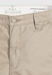 Pantaloni chino din bumbac bej, cu o croială dreaptă, dotati cu închidere frontală cu buton, bucle pentru curea și detalii cusute pe cusături și buzunare.