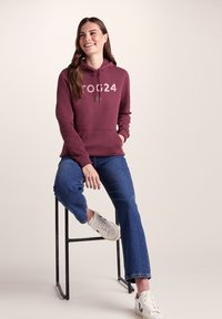 Woman sitting on a stool wearing a burgundy TOG24 hoodie, blue jeans, and white sneakers, smiling and looking to the side.