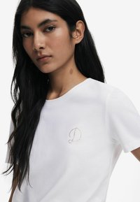 Weißes Baumwoll-T-Shirt mit kurzen Ärmeln, das auf der linken Brustseite ein kleines, goldakzentuiertes 'D'-Logo im Perlen-Design zeigt.