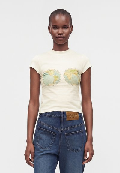 Γυναίκα με κοντά μαλλιά που φοράει λευκό t-shirt με εκτυπώσεις παγκόσμιου χάρτη στο στήθος και μπλε τζιν με καφέ δερμάτινη ετικέτα.