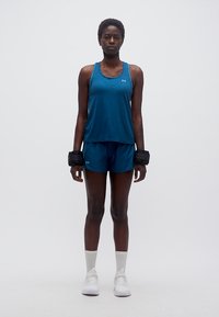 Αθλητική γυναίκα στέκεται με το πρόσωπο προς τα εμπρός, φορώντας μπλε αμάνικο μπλουζάκι και σορτς, λευκά αθλητικά παπούτσια και βαρίδια καρπού, σε απλό λευκό φόντο.