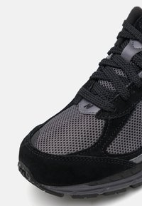 Musta ja harmaa lenkkari, jossa verkkoyläosa, mokkanahkaiset yksityiskohdat, teksturoitu pohja ja litteät nauhat; sivussa logo ja pehmustettu kaulus.