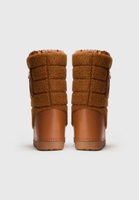 Brūni viduslīdz ceļiem ziemas zābaki ar quilted auduma augšdaļu un gludas ādas segtu papēdi, skatīti no aizmugures uz baltas fona.