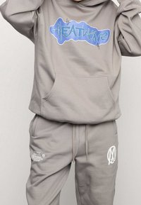 Persona vestida con un hoodie gris claro y joggers con diseños en azul y blanco de "Heatwave" y logotipo, posando con las manos cerca de la cabeza.