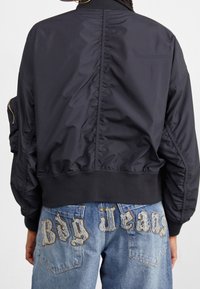 Zwarte nylon bomberjack met geribde manchetten, met gerimpelde details op de rug. Draagt bij blauwe denim jeans, versierd met "Big Jeans" borduursel.