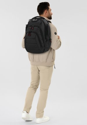 Man in beige outfit wearing and holding a large black backpack with multiple zippered compartments, standing sideways on a white background.