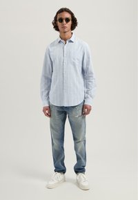 Lys blå stripete skjorte med knapper og brystlomme, kombinert med slitte lyseblå jeans og hvite joggesko. Sirkulære mørke solbriller.