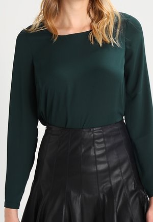 Vrouw draagt een donkergroene blouse met lange mouwen, ingestopt in een zwarte geplooide leren rok met hoge taille, afgebeeld van nek tot midden dij.