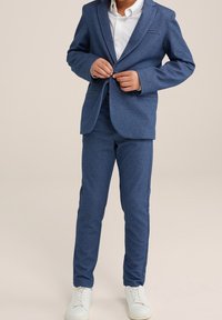 Completo blu realizzato in tessuto testurizzato, composto da un blazer e pantaloni aderenti, abbinato a una camicia bianca e sneakers bianche.