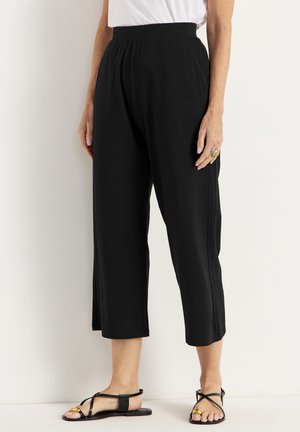 Zwarte cropped broek met een gladde textuur, elastische taille en zijsplitsingen, gecombineerd met zwarte sandaaltjes met banden op een neutrale achtergrond.