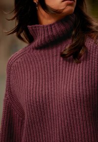 Strickpullover in tiefem Pflaumenton mit hohem Kragen und geripptem Textur. Der lockere Schnitt betont weite Ärmel und ein kräftiges Design.