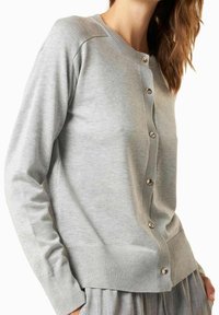 Cardigan gris clair avec encolure ronde, boutons métalliques sur le devant, poignets et ourlet côtelés, fabriqué en tissu doux et texturé.