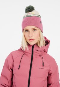 Rosa isolierte Jacke mit hohem Kragen und schwarzem Reißverschluss. Begleitet von einer hellrosa Mütze mit einem grünen Pompon und einem Skischriftzug.