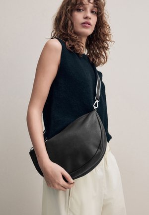 Frau mit lockigem Haar, die ein ärmelloses schwarzes Oberteil und helle Hosen trägt, hält eine große schwarze Lederschultertasche.