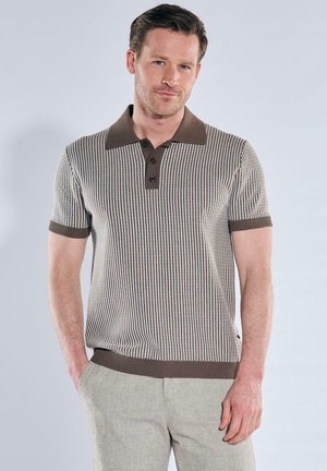 Mann mit kurzem braunem Haar, der ein braun-weiß gestreiftes kurzärmeliges Poloshirt und hellbeige Hosen trägt, stehen mit einer Hand in der Hosentasche.