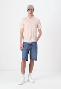 Světle růžové polo tričko na zip s krátkým rukávem, doplněné džínovými šortkami. Model má na sobě béžovou čepici a bílé tenisky. Jednoduchý design, ležérní střih.