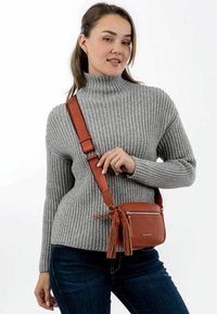 Borsa a tracolla in pelle marrone con scomparto con zip e dettagli con frange. Indossata con un maglione grigio a coste e jeans blu.