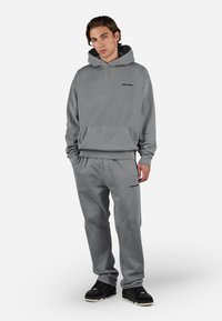 Conjunto de sudadera con capucha y pantalones deportivos grises, hecho de tela suave, con un bolsillo delantero y acentos de logo negro. El modelo lleva zapatillas negras.