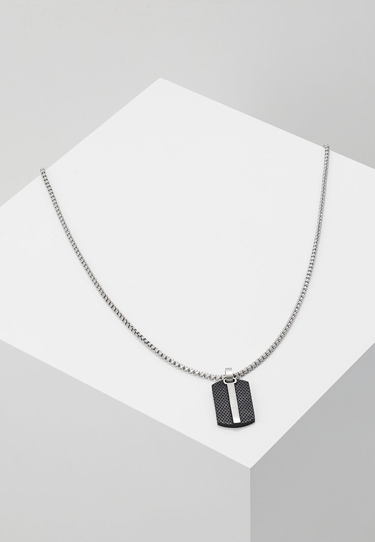 Collier chaîne en argent avec un pendentif plaque militaire noir rectangulaire, doté d'une bande argentée verticale, présenté sur une surface géométrique blanche.