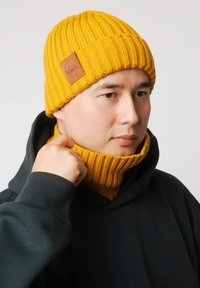 Żółta dzianinowa czapka z prążkowaną fakturą i skórzaną naszywką, noszona z ciemną bluzą z kapturem o wysokim kołnierzu, dopasowanym do koloru czapki.