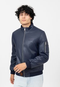 Veste bombardier en cuir bleu marine avec un col haut, fermeture éclair et poignets côtelés. Comprend une poche à patch sur la manche gauche et un logo subtile.