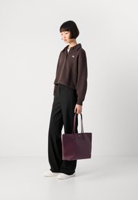 Brown cropped sweatshirt with a collar and logo, paired with black wide-leg pants. Holding a purple tote bag, wearing white sneakers.