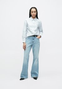 Cămașă cu dungi albastre, cu nasturi și guler, asortată cu blugi evazați deschiși la culoare. Pantofi negri cu vârf ascuțit completează ținuta.