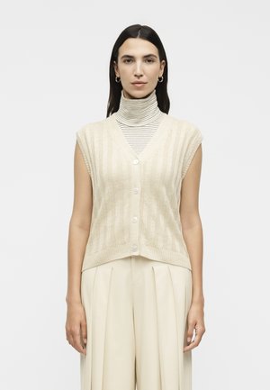 Γυναίκα με ίσια σκούρα μαλλιά που φοράει κρίκους σκουλαρίκια, μια μπεζ αμάνικη γιλέκο με κουμπιά πάνω από ένα ριγέ ζιβάγκο και ανοιχτόχρωμο πιετέ παντελόνι.