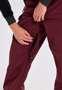 Bordeauxfärgade utomhusbyxor med dragkedjeförsedda sidofickor, tillverkade av mjukt, texturerat tyg. Justerbart hårdvarudetalj för en säker passform.