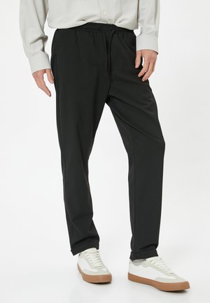 Svarta byxor med resårmidja och dragsko, med sidofickor och avsmalnande passform, kombinerade med vita och grå sneakers.