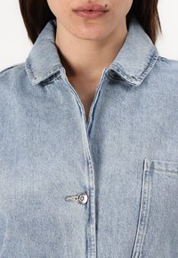 Giacca in denim blu chiaro con colletto, chiusura con un solo bottone e tasca frontale con dettagli di cucitura visibili. La texture appare leggermente sbiadita.